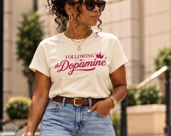 Könnte beinhalten: Ein cremefarbenes Kurzarm-T-Shirt mit dem Text FOLLOWING the Dopamine und einer kleinen roten Krone, kombiniert mit einer hellblauen weiten Jeans, einem braunen Ledergürtel, einer leuchtend pinken geflochtenen Handtasche und passenden Pumps.