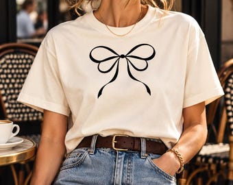 Könnte beinhalten: Ein cremefarbenes Kurzarm-T-Shirt mit einer mittig platzierten schwarzen Grafik einer Schleife. Das Oberteil wird mit einer hellblauen Jeans, einem braunen Ledergürtel mit goldener Schnalle und einer dunkelbraunen geflochtenen Handtasche kombiniert.