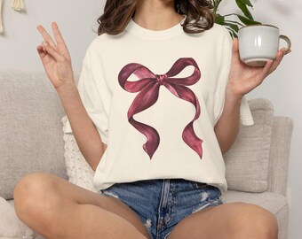 Könnte beinhalten: Ein cremefarbenes Kurzarm-T-Shirt mit einer großen, mittigen Grafik einer dunkelburgunderroten Schleife. Das T-Shirt hat einen Rundhalsausschnitt und eine lockere Passform und zeigt eine detaillierte Illustration der Stofffalten und Schatten.