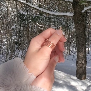 May include: A close-up of a hand wearing a stack of five thin copper rings on the thumb. The rings are a warm, reddish-brown color and have a slightly textured surface. The hand is in focus, while the background is blurred and shows a snowy forest scene.