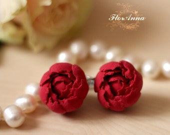 May include: A pair of red polymer clay earrings shaped like peonies. The earrings are displayed on a white surface with a pearl necklace.