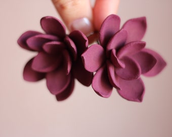 May include: Two burgundy-colored flower-shaped hair accessories. Each flower has multiple layered petals, creating a textured appearance. The background is a soft, neutral tone, highlighting the deep color of the floral accents. These are likely hair clips or barrettes.