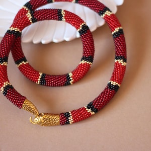 Puede incluir: Un collar de cuentas con un diseño tubular flexible y un patrón repetitivo de cuentas rojas, negras y doradas. El collar termina en un cierre de cabeza de serpiente de metal dorado texturizado que se engancha en el extremo opuesto del hilo.