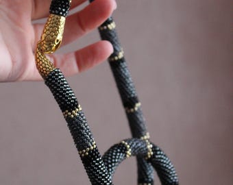 Puede incluir: Un collar de serpiente de cuentas negras y doradas con un dije de cabeza de serpiente dorada.