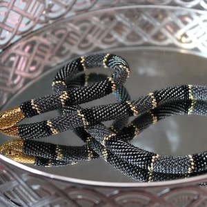 May include: A black and gold beaded necklace in the shape of a snake. The necklace is coiled and resting on a silver tray with a decorative pattern.