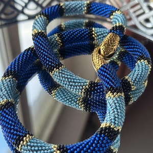 Op de afbeelding: Een kralen slangenarmband in blauwe, zwarte en gouden tinten. De armband is opgerold, met een goudkleurige slangenkopsluiting. De kralen zijn klein en strak geweven, waardoor een textuur ontstaat. De armband wordt op een reflecterend oppervlak getoond.