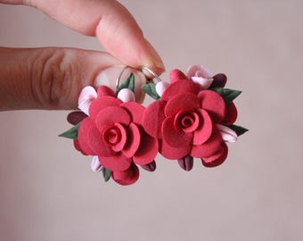 May include: A pair of handmade earrings featuring vibrant red rose-shaped designs. Each earring showcases detailed petals, complemented by green leaves and small, light pink accents. The earrings are held up against a soft, neutral background.