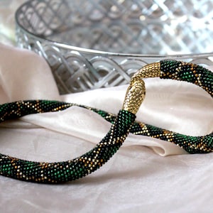 Op de afbeelding: Een groene en gouden kralen slangenketting. De ketting is in een cirkel gekruld en heeft een gouden kop.