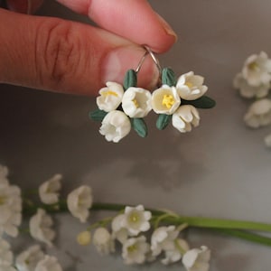Op de afbeelding: Witte bloemen oorbellen met gele centra en groene bladeren. De oorbellen worden vastgehouden door de vingers van een persoon. De oorbellen zijn gemaakt van kleine witte bloemen met gele centra en groene bladeren. De oorbellen zitten aan een zilveren hoepel.