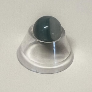May include: A dark green and gray gemstone sphere sits atop a clear, conical display stand. The sphere is divided into two distinct color sections. The stand has a wide, circular base and a flared shape.