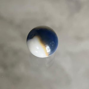 May include: A glass marble with a white base, transitioning to a dark blue and brown swirl. The marble is round and has a glossy finish. This collectible toy is suitable for games or display.