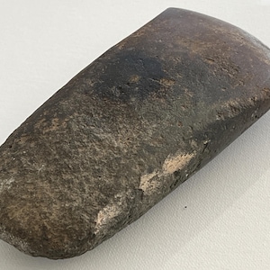 May include: A weathered, hand-shaped stone axe head. The axe head is a dark, mottled brown with a smooth, polished edge and a rough, textured surface. The axe head is a prehistoric tool.