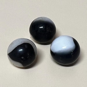 May include: Three glass marbles with a black and white swirl design. The marbles are spherical and have a glossy finish. The color pattern varies on each marble, with some having more black or white.