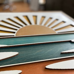 May include: A wooden wall art piece depicting a sunset over the ocean. The sun is made of layered wood with a brown center and white rays. The ocean is represented by a teal blue layer of wood with white waves layered on top.