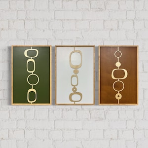 May include: Three framed wood wall art pieces with a mid-century modern design. Each piece features a different color background: green, white, and brown. The design is a series of gold circles and ovals in a vertical line.