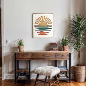 May include: A framed print of a retro sunset over a body of water with a yellow sun and orange, red, green, and blue stripes. The print is hanging on a white wall above a wooden desk with a white furry stool.