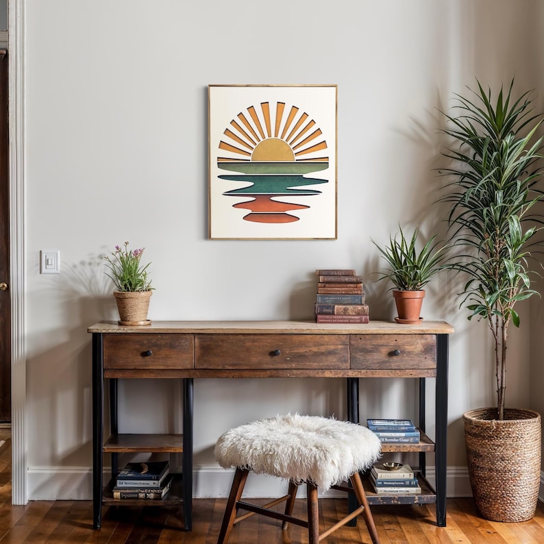 May include: A framed print of a retro sunset over a body of water with a yellow sun and orange, red, green, and blue stripes. The print is hanging on a white wall above a wooden desk with a white furry stool.