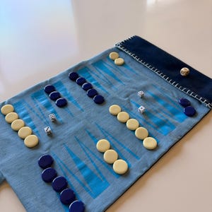May include: A backgammon game set on a light blue playing surface with a dark blue border. The set includes ivory and dark blue game pieces, and dice. The game is set up on a white table.