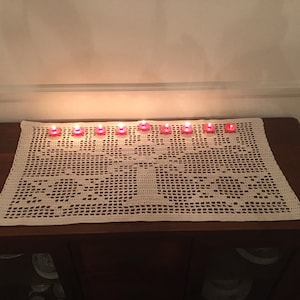Puede incluir: Un camino de mesa de crochet blanco con un diseño floral, decorado con 10 pequeñas velas rojas.