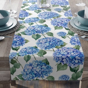 Pode incluir: Um corredor de mesa branco com um padrão floral azul e verde. O padrão apresenta grandes flores de hortênsia azuis com folhas verdes.