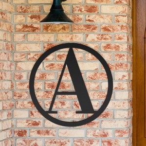 May include: Black metal letter A in a circle, mounted on a brick wall. The letter is in a modern, minimalist style.