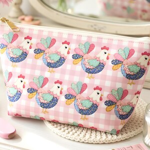 May include: A pink and white gingham cosmetic bag with a repeating pattern of colorful rooster illustrations. The bag has a gold zipper and a white interior. The rooster design features patchwork-style details in various colors.