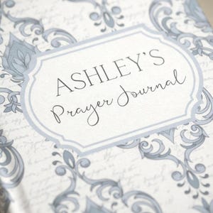 Könnte beinhalten: Ein Hardcover-Journal mit einem hellblauen und weißen Blumenmuster. Auf dem Einband steht in dekorativer Schrift der Text "ASHLEY'S Prayer Journal", eingerahmt von einem hellblauen Rand. Der Hintergrund hat ein subtiles Schriftmuster.