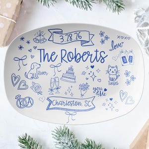 May include: A white ceramic platter with blue hand-drawn illustrations and text. The platter features the text "The Roberts", the date "3.28.26", and the location "Charleston, SC". Other drawings include a dog, cat, cake, and hearts.