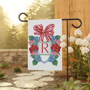May include: A white garden flag with a red, white, and blue bow, red poppies, blue hydrangeas, and American flags surrounding a blue and white shield with the letter "R" in red.