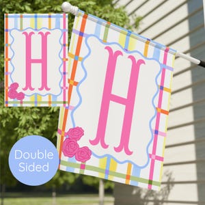 Puede incluir: Bandera de jardín de doble cara con una gran letra rosa "H" sobre un fondo blanco. La bandera tiene un borde azul festoneado y rosas rosas. El fondo presenta un patrón de cuadros colorido.