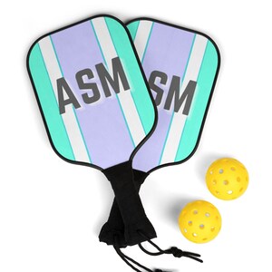 Könnte beinhalten: Zwei Pickleball-Schläger mit einem türkis-, lavendel- und weiß gestreiften Design und den Buchstaben "ASM" in fetter schwarzer Schrift. Zwei gelbe, perforierte Pickleballs sind ebenfalls enthalten. Die Schläger haben schwarze Griffe.