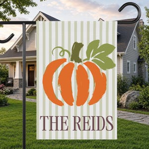 May include: A rectangular garden flag featuring a vertical light green and white striped background. Centered is a large orange pumpkin illustration with a green stem and leaf. The text THE REIDS is printed in dark purple at the bottom.