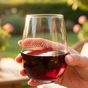 Può includere: Un bicchiere da vino senza stelo trasparente pieno di vino rosso, tenuto da una persona. Il bicchiere ha le parole "chapter 50" incise sul lato. Lo sfondo è sfocato, mostrando vegetazione e luce solare.
