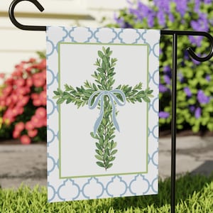 May include: A garden flag featuring a cross design made of green leaves and a light blue ribbon. The cross is centered on a white background with a green and blue border and a blue quatrefoil pattern.