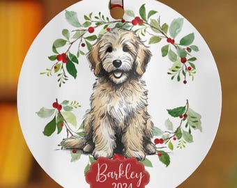 May include: A white round Christmas ornament with a watercolor illustration of a golden doodle puppy sitting in a wreath of green leaves and red berries. The ornament has the name "Barkley" and the year "2024" printed on it.