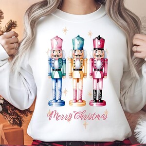 Puede incluir: Sudadera blanca con una ilustración de acuarela de tres cascanueces en verde, amarillo y rosa. El texto "Merry Christmas" está impreso debajo de los cascanueces.
