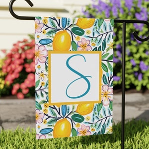May include: A white garden flag with a floral pattern featuring lemons, leaves, and pink flowers. The letter "S" is in a turquoise blue color and is centered on the flag. The flag is hanging on a black metal pole.