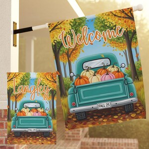 May include: A teal pickup truck filled with pumpkins is the focal point of this fall-themed garden flag. The flag reads "Welcome" in orange script, with a backdrop of autumn trees. A smaller flag shows the name "Langley".