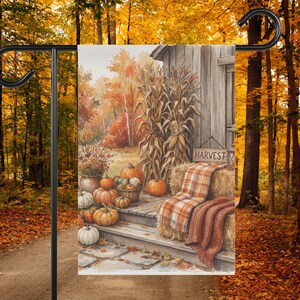 Puede incluir: Una bandera de jardín rectangular con una ilustración en acuarela de una escena de porche rústico con calabazas, tallos de maíz y balas de heno. Un letrero dice HARVEST. La escena incluye una manta a cuadros sobre una bala de heno.