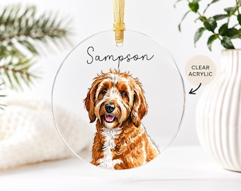 May include: A round clear acrylic ornament featuring a colorful illustration of a brown and white dog with the name Sampson printed in black script above the dog. The ornament hangs from a gold ribbon loop at the top center of the circular piece.