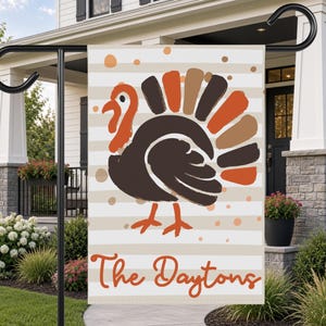 May include: A rectangular garden flag featuring a stylised turkey illustration in shades of brown and orange against a background of horizontal beige stripes and scattered dots. The text The Daytons is printed in orange script at the bottom.