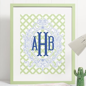 May include: Framed print featuring a blue monogram within a decorative wreath on a light green trellis pattern background. The frame is a matching light green. A desk lamp and cactus figurine are visible.