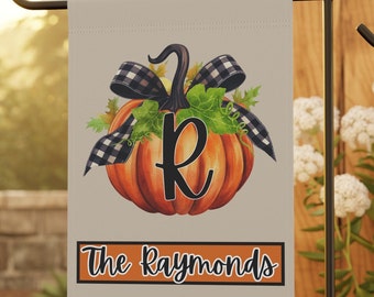 Könnte beinhalten: Beige Gartenflagge mit einem Aquarell-Kürbis, einer schwarz-weiß karierten Schleife und grünen Blättern. Der Kürbis hat einen schwarzen Buchstaben "R" und den Schriftzug "The Raymonds" auf einem schwarzen und orangefarbenen Banner.