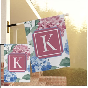 Könnte beinhalten: Eine weiße Gartenflagge mit einem rosa-blauen Blumenmuster. Die Flagge zeigt ein großes, rotes Quadrat mit einem weißen Buchstaben "K" in der Mitte. Die Flagge ist perfekt, um Ihrem Außenbereich einen Hauch von Eleganz zu verleihen.