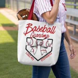 Op de afbeelding: Een witte canvas draagtas met rode hengsels voorzien van de tekst Baseball Grandma in rode schrijfletters. Onder de tekst staat een afbeelding van een honkbal honkplaat met een grote strik van stof met een honkbalpatroon.