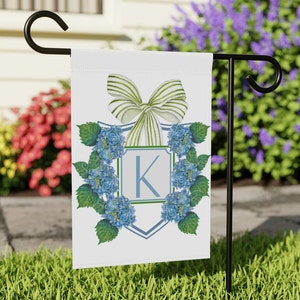May include: A white garden flag with a blue and green monogram design. The monogram features the letter "K" surrounded by blue hydrangeas and green leaves. A green and white striped bow is at the top of the design.