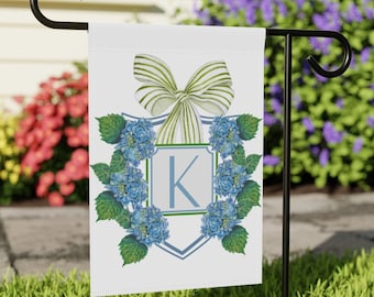 May include: A white garden flag featuring a blue and green monogram design. The monogram displays the letter "K" encircled by blue hydrangeas and green foliage. A green and white striped bow sits atop the design.