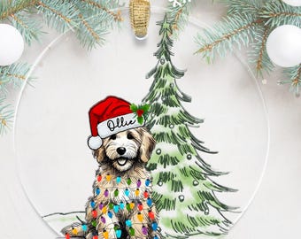 May include: A personalized Christmas ornament featuring a doodle dog wearing a Santa hat and tangled in Christmas lights, with a green Christmas tree in the background. The ornament is clear and round, with the name "Ollie" written on the dog's hat.