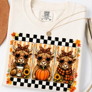 May include: A cream-colored t-shirt with a Comfort Colors label featuring a graphic of three Highland cows wearing heart-shaped sunglasses and plaid headbands. The cows are holding a coffee cup, a pumpkin, and wearing a plaid scarf with sunflowers.