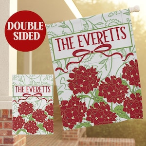 Op de afbeelding: Dubbelzijdige tuinvaandel met de naam "The Everetts" in rood schrift boven een rode strik en rode hortensiabloemen. De vlag heeft een witte achtergrond met groene bloemaccenten. Een kleinere versie van de vlag is ook zichtbaar.
